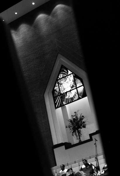 The church choir sit below a stained glass with the holy white dove above it.  